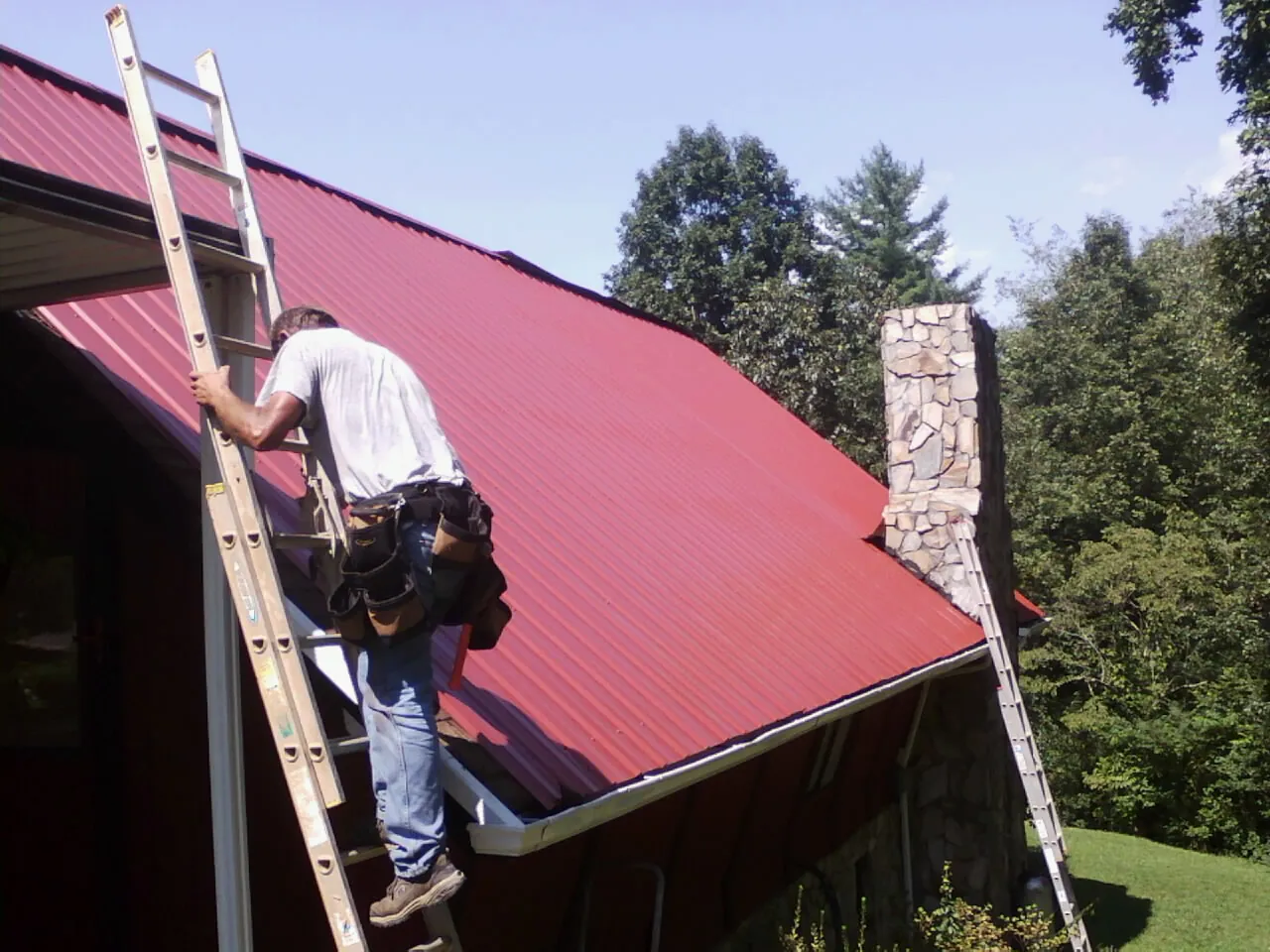 Roofer on a ladder beside a red metal roof and stone chimney — Soffit & Fascia Repair by Roofing Brothers in Arvada